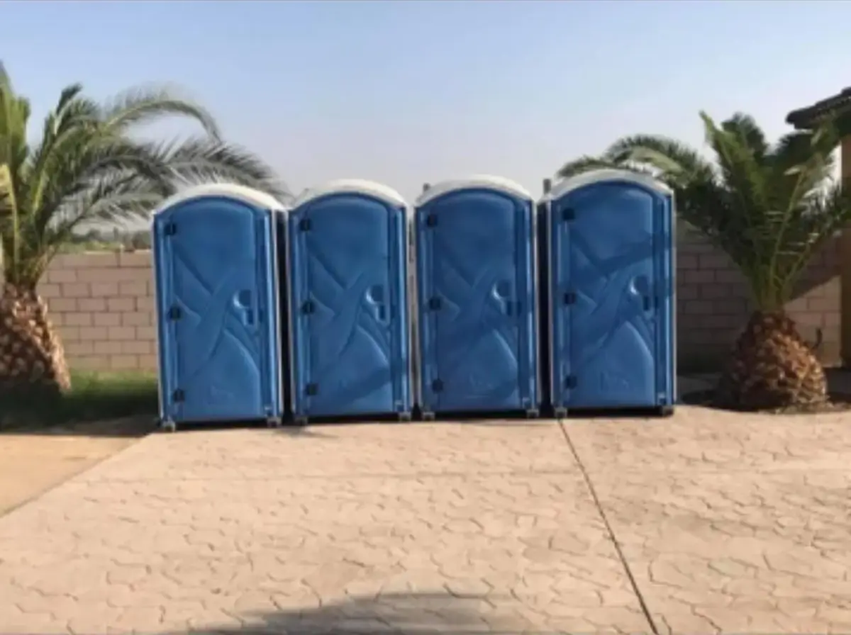 Four blue standard porta potties lined up between palm trees